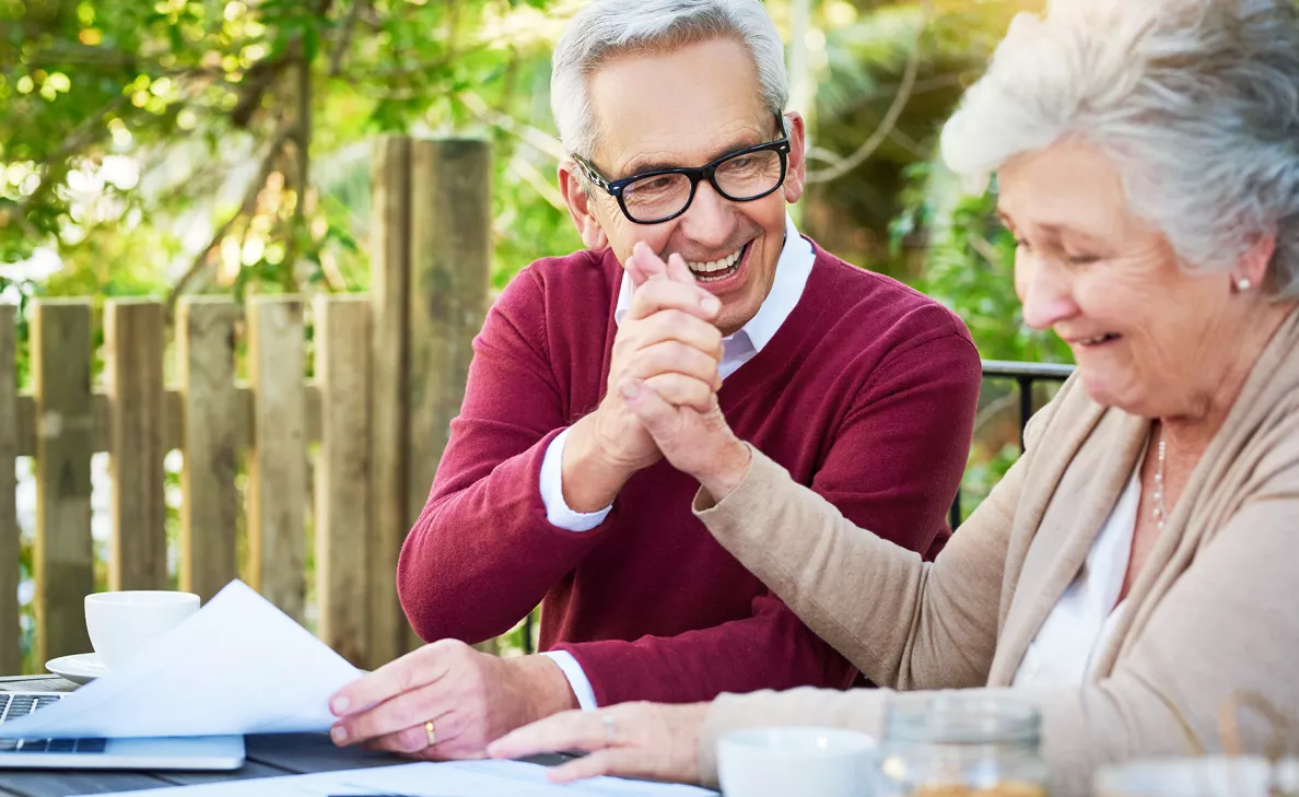  A retirement-aged couple smile and hold hands as they look at documents on their porch.

