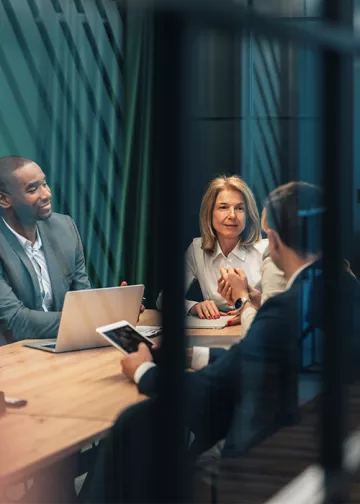  Three professionals sit around a wooden conference table in a modern office, engaged in discussion, with a laptop and smartphone visible; the scene is partially framed by glass or architectural elements in the foreground.
