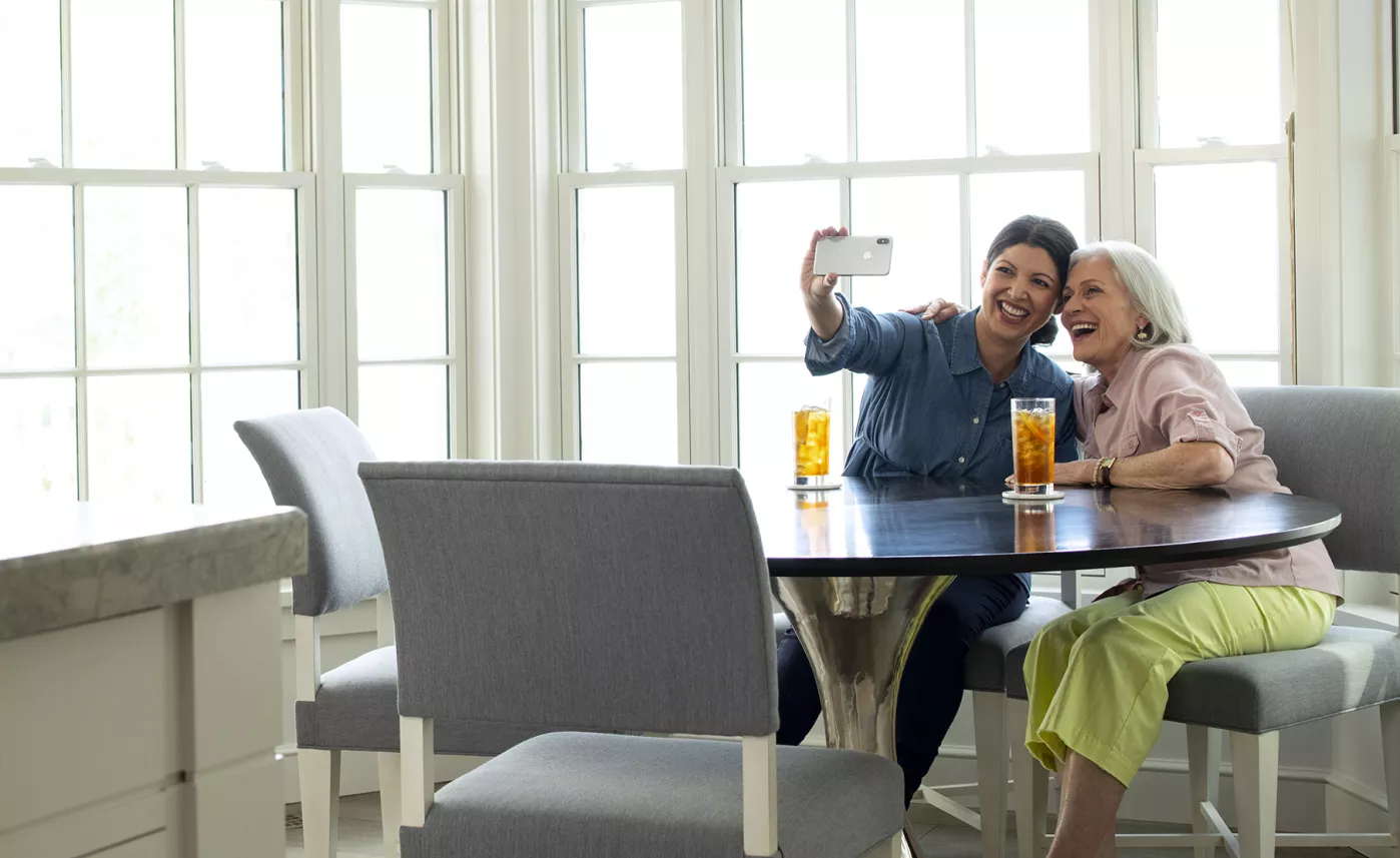  A woman and her retirement-aged mother pose for a selfie at their kitchen table.
