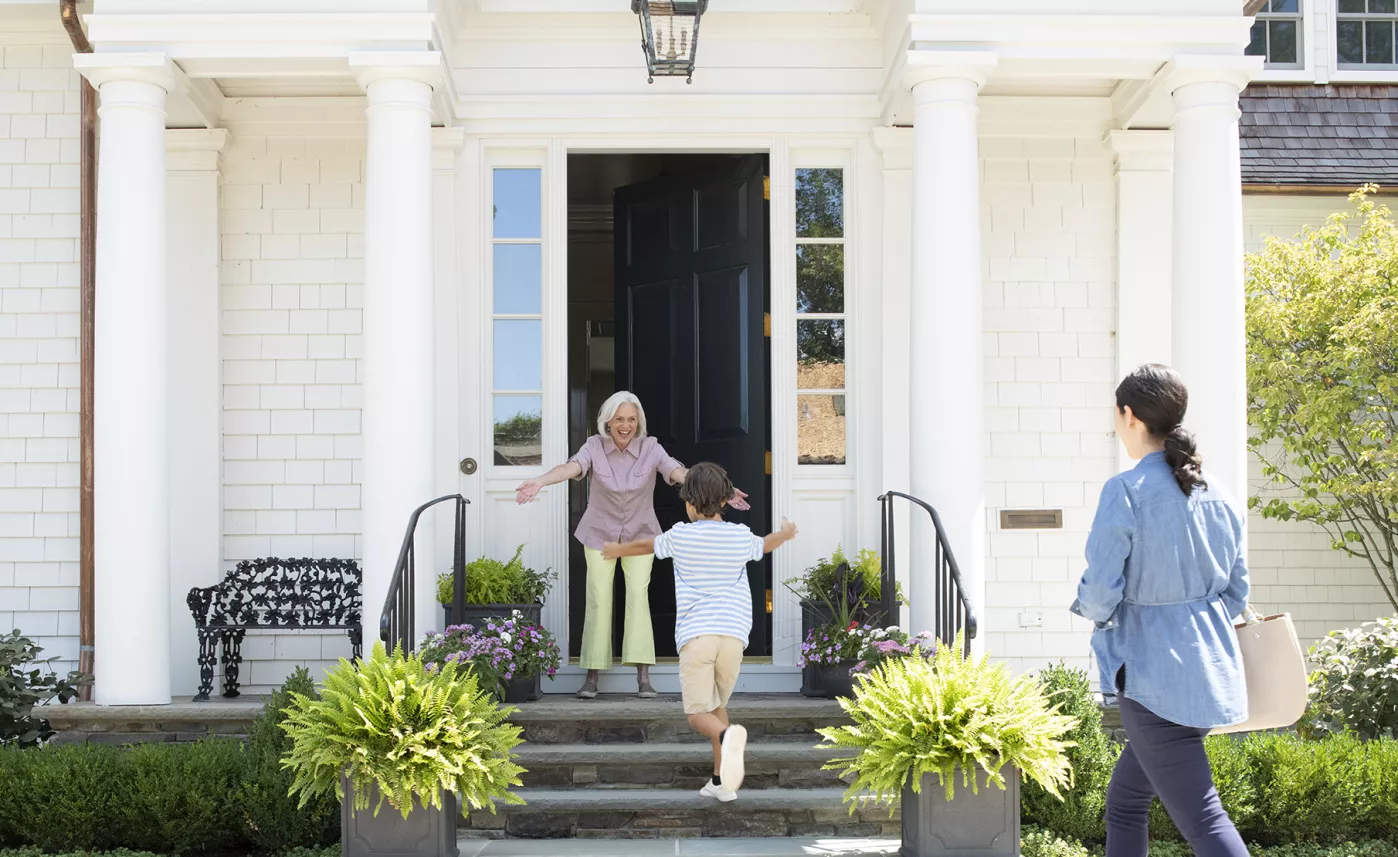  A young child runs up the front steps of a large house to greet his grandmother as his mom watches and smiles.

