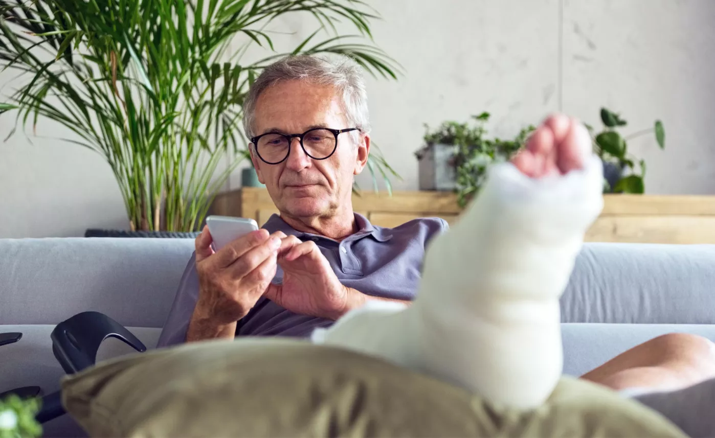  Man with cast on foot sitting on sofa using his smartphone.
