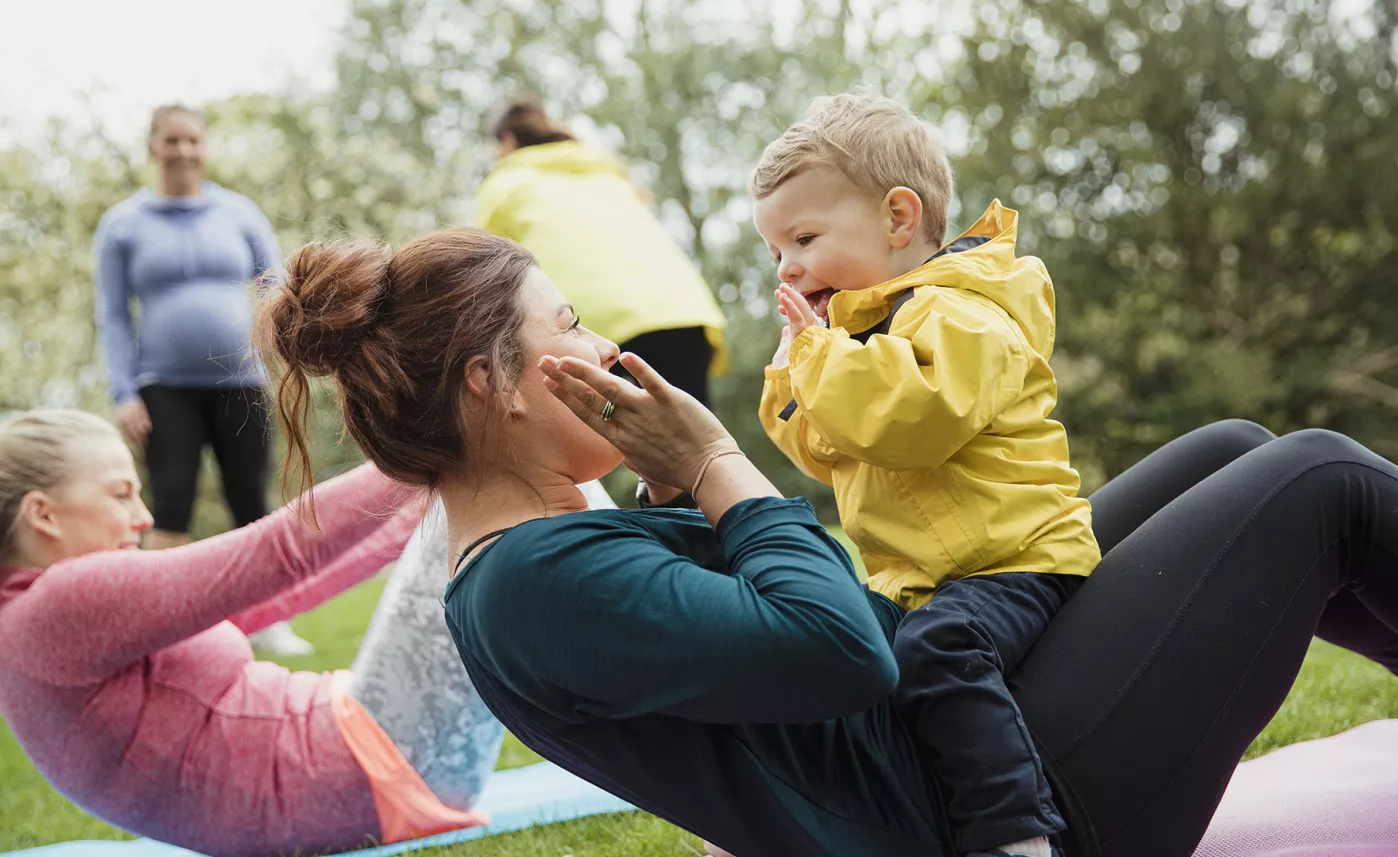  A young mother exercises at a park with her toddler son and they smile at one another.
