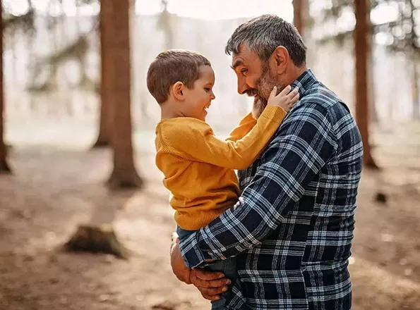  A retirement-aged man holds his young grandson during a hike on a trail through the woods.

