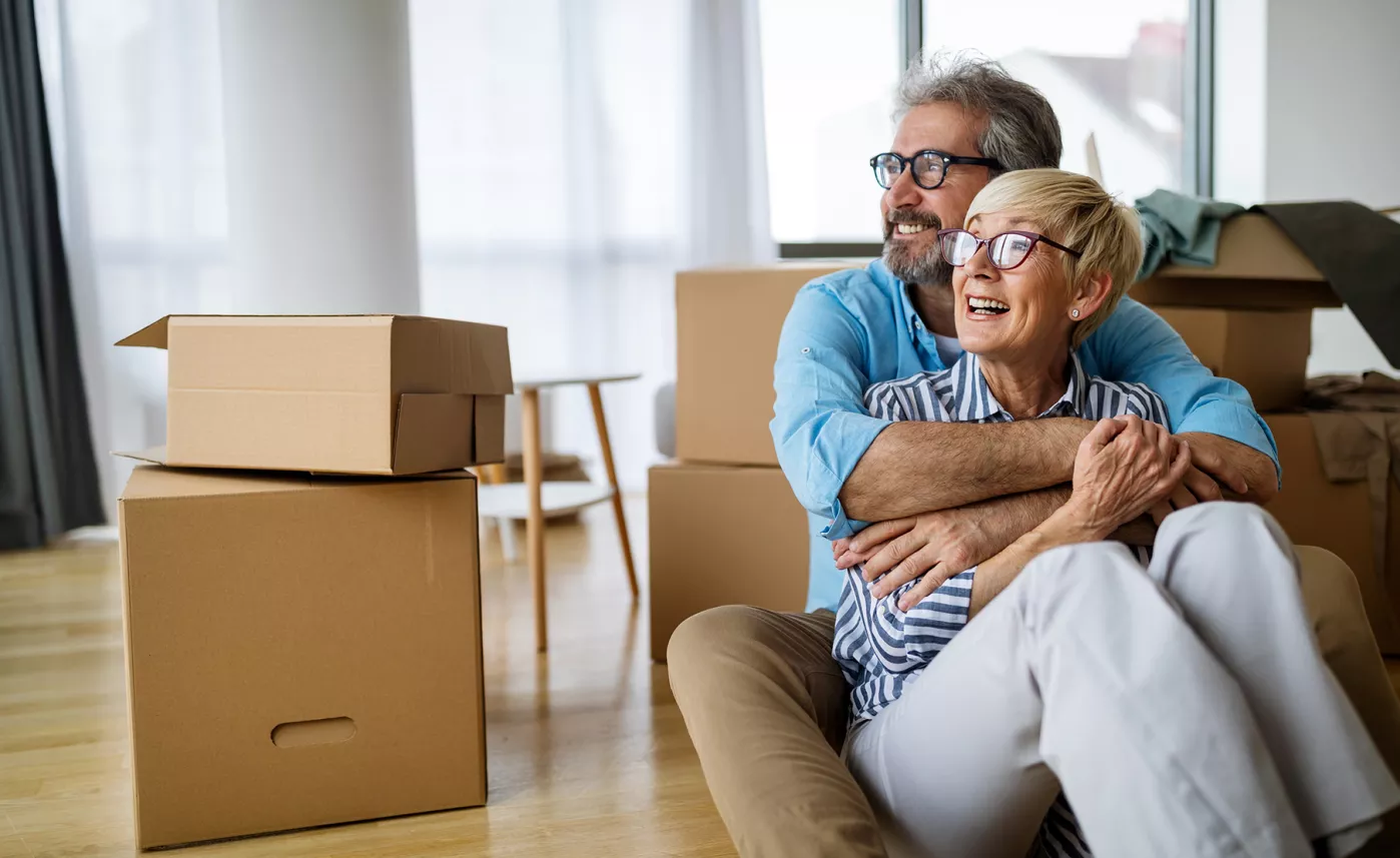  A smiling retirement couple sit on the floor amongst moving boxes

