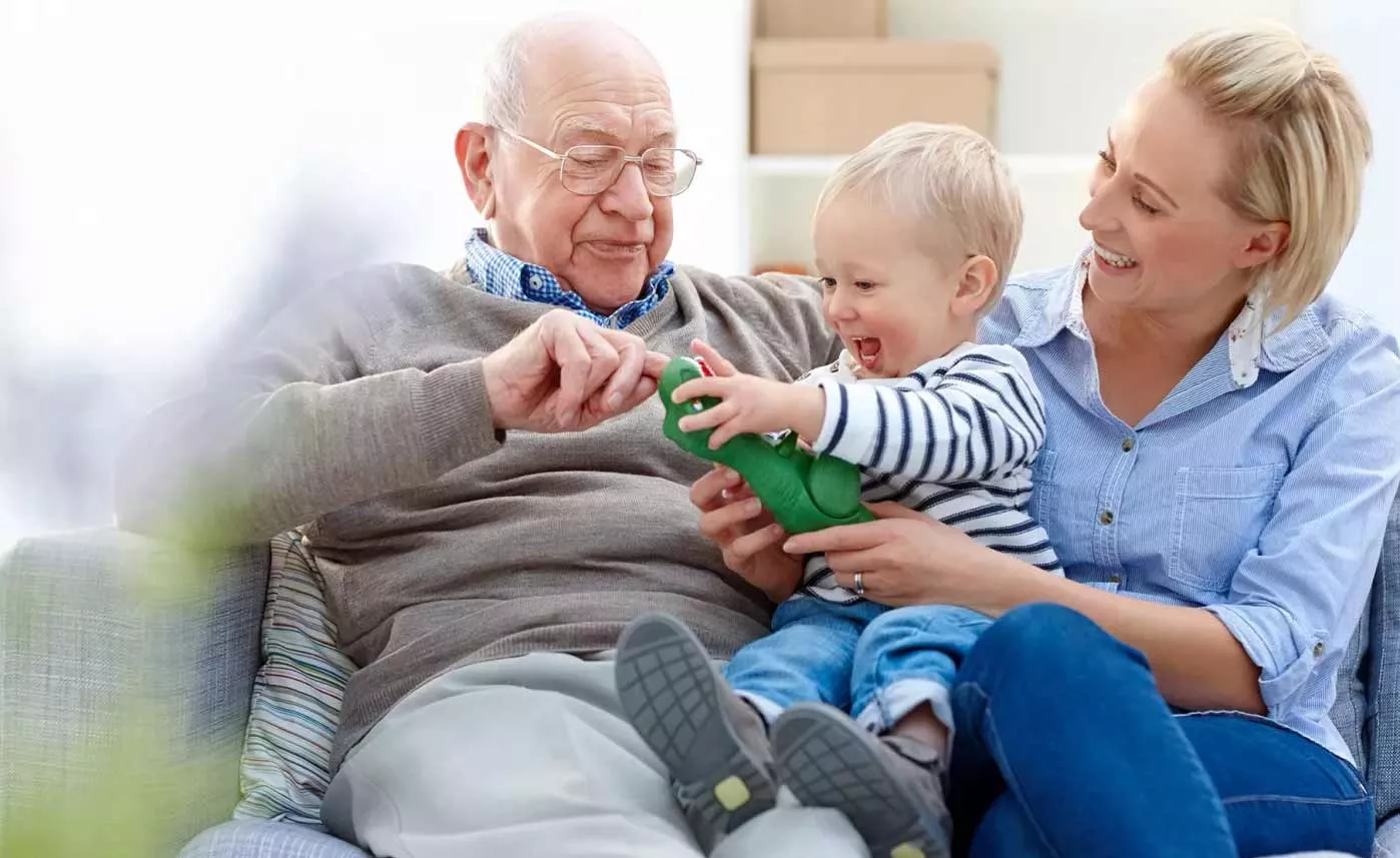  An older man sits on his couch with his adult daughter and his baby grandson.
