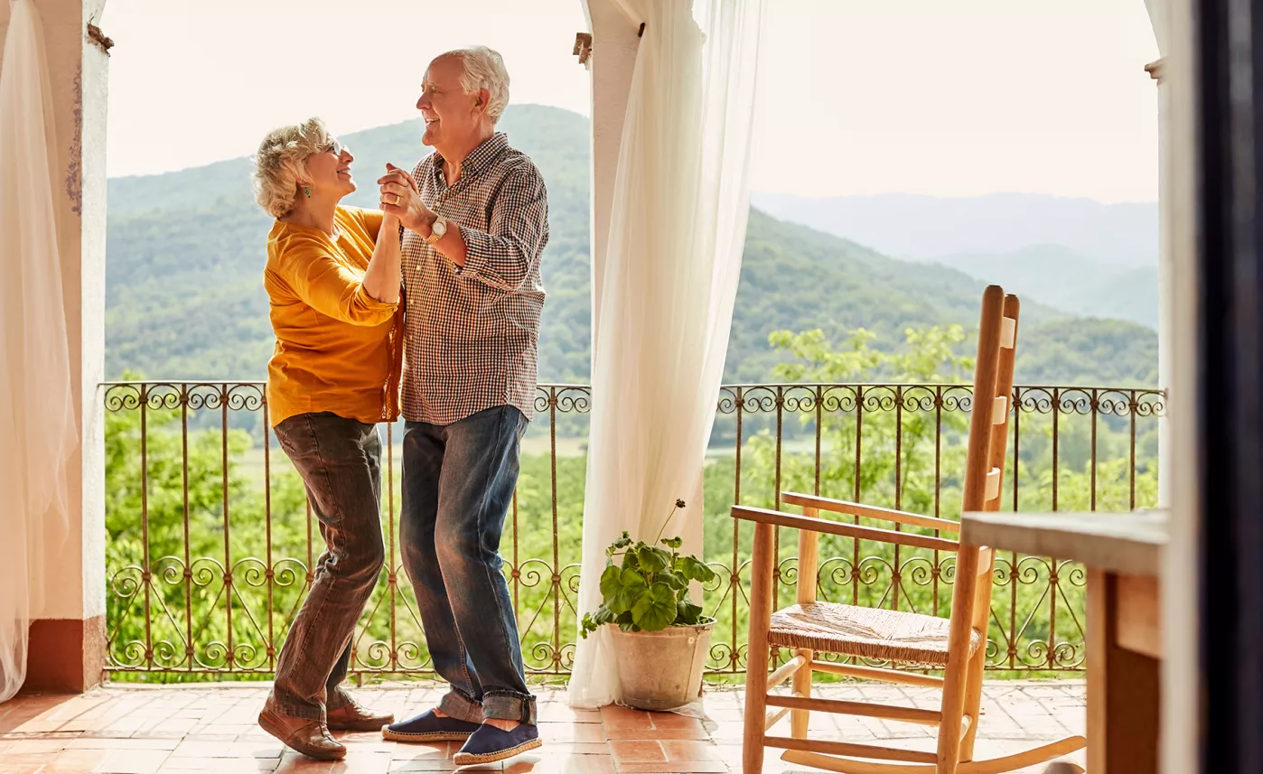  A retirement-aged couple dances together on a porch with a view of mountains.

