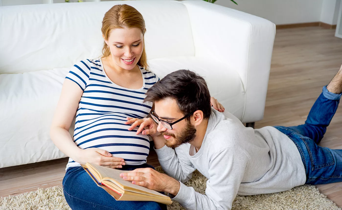  A young pregnant couple sit in their living room and read from a book together.
