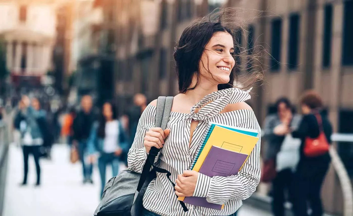  A college student walks through a crowded campus, smiling and carrying notebooks and a book bag.
