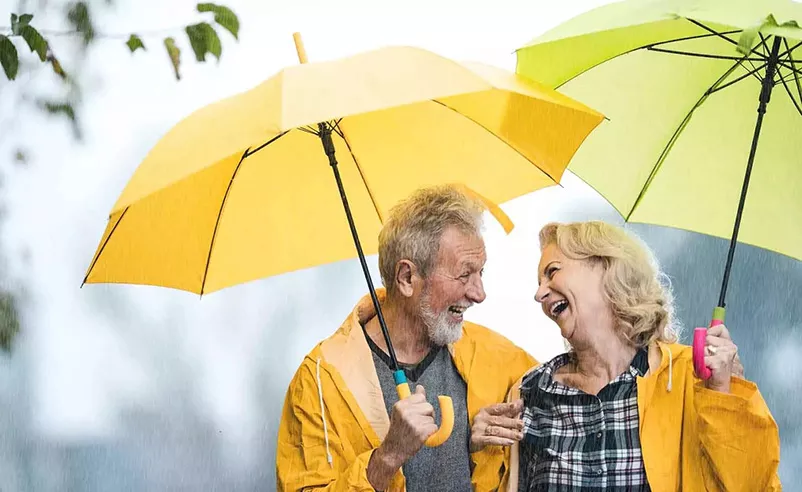 A senior couple laughs in the rain, with umbrellas and bright yellow raincoats, and enjoy the moment.