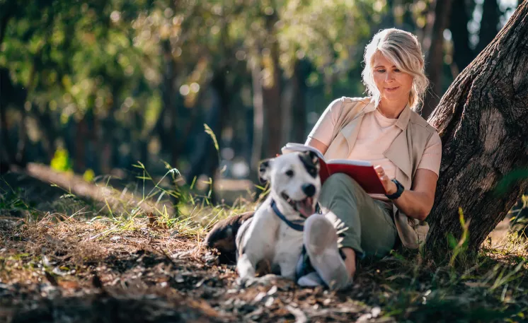 Happy mature woman sitting under a tree in the woods with her dog while reading a book.