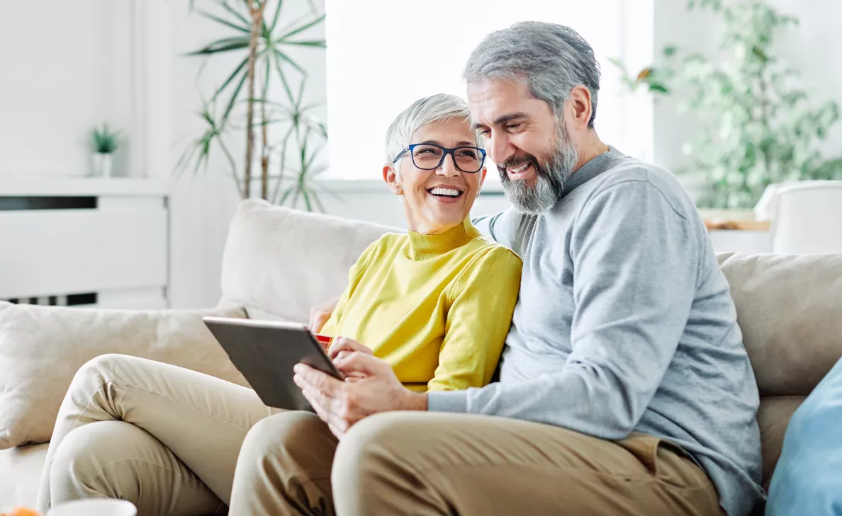 A retirement-age couple smiles as they read from a tablet on the couch.
