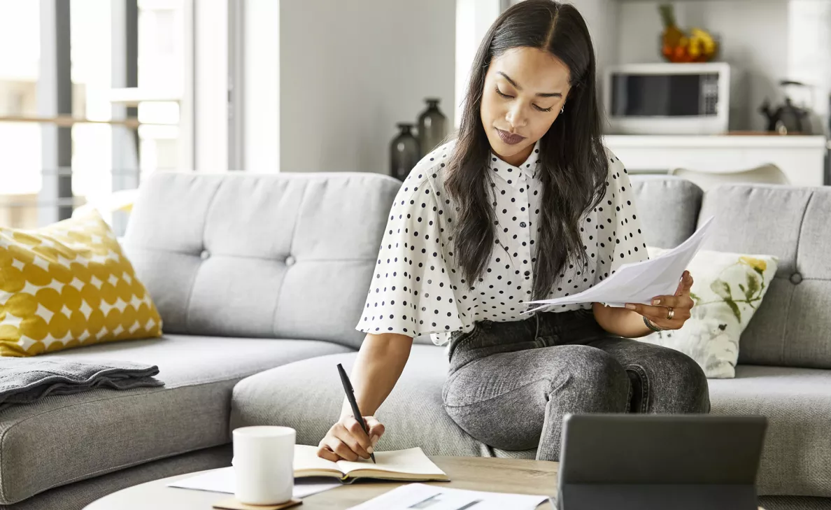 A woman reads from a printed document and takes notes in a notebook.