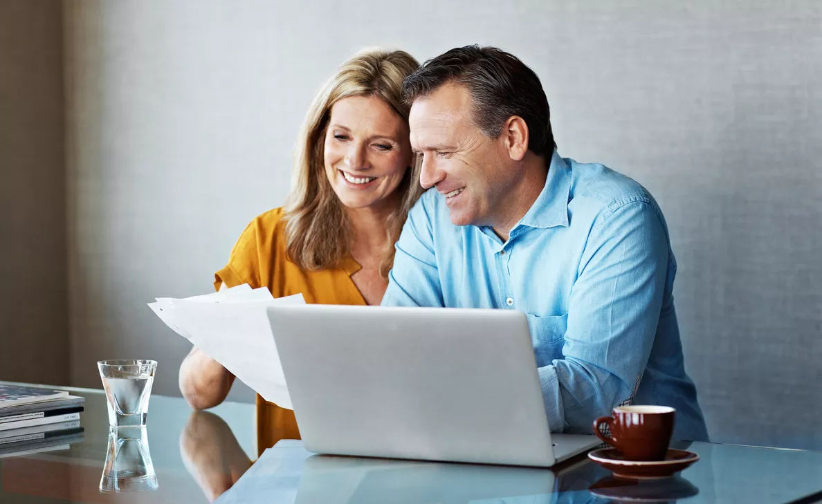 A middle-aged couple smiles as they review printed tax documents in front of their laptop.