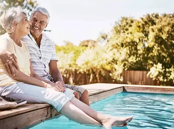A retirement-aged couple smile together as they dip their feet in their pool on a sunny day.