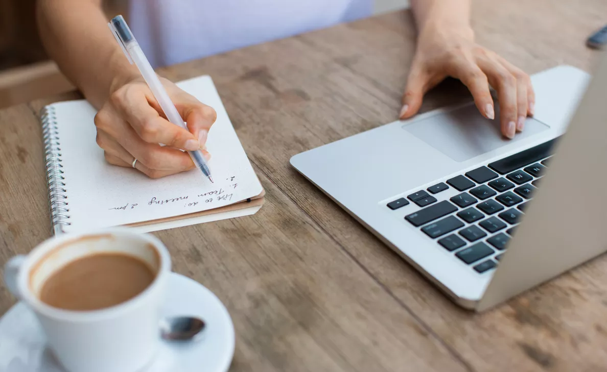 A woman writes in a notebook while scanning her laptop.