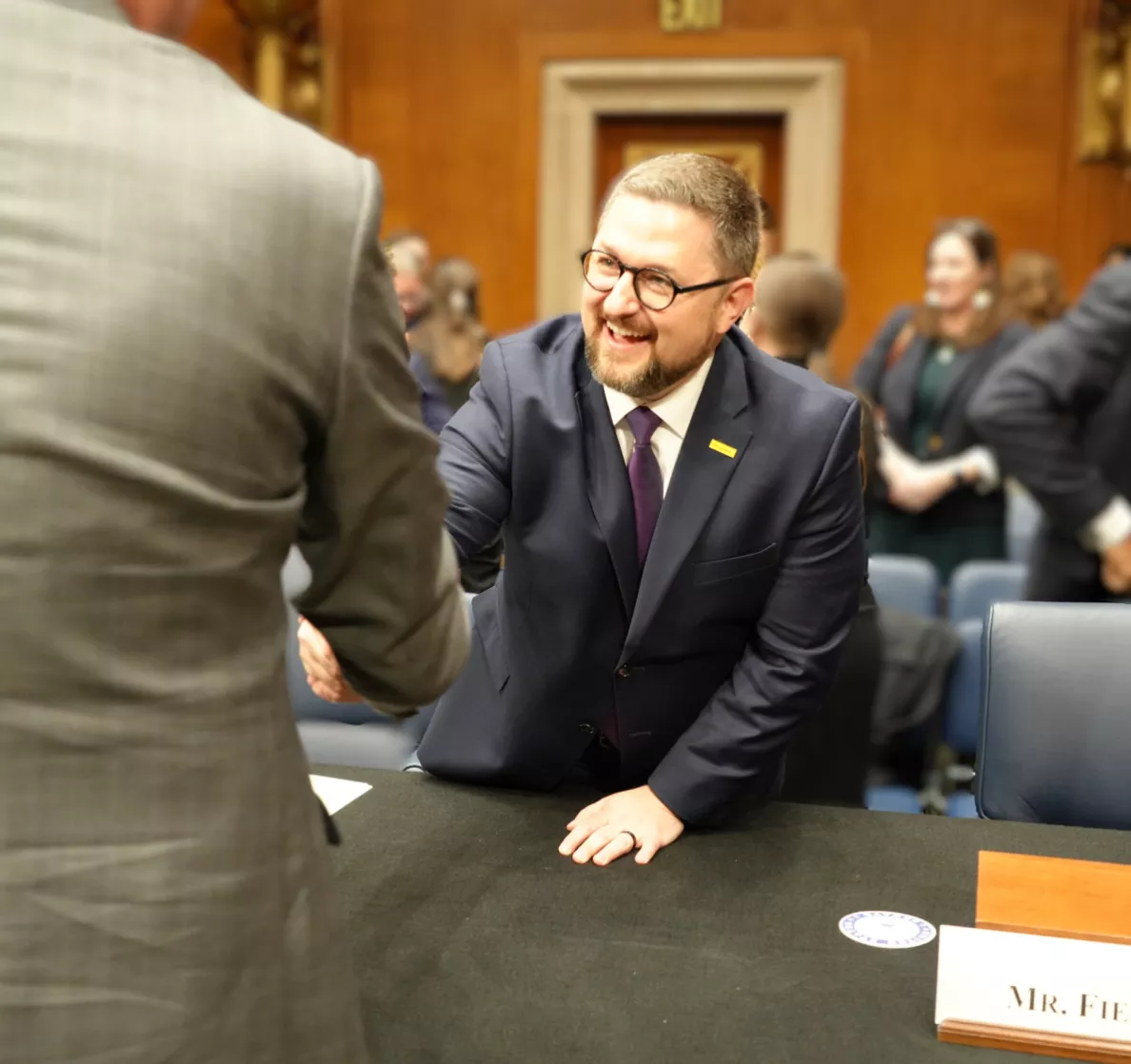  Person at a hearing room table shaking hands with another person, with a nameplate reading ‘Mr. Fiesta’ in front.
