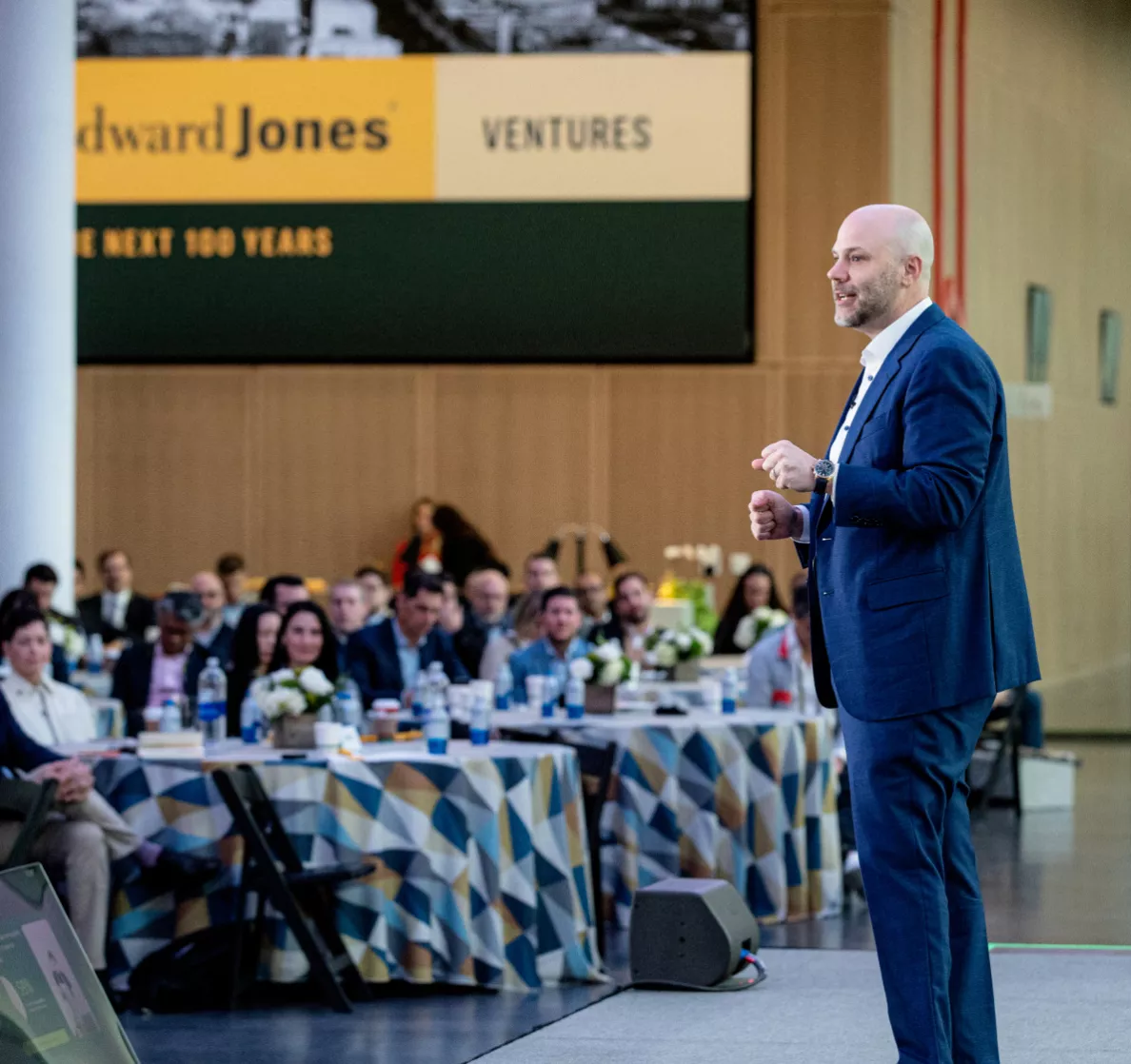  A person in a suit speaking on stage in front of an audience at a corporate event.
