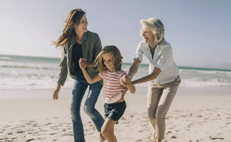 A retirement-aged woman laughs on the beach with her adult daughter and young granddaughter.