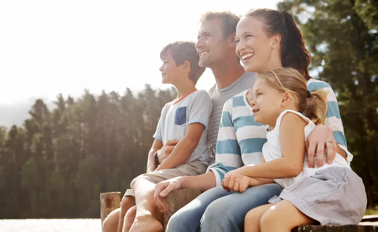 A young family sits together on a dock and smiles at the view of a lake. A young family sits together on a dock and smiles at the view of a lake.