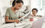 A young mother goes through financial records at the kitchen table as her toddler son looks on.