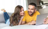 A married couple sits on their couch and reads about financial tips from their laptop.