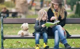A young mother sits with her toddler daughter on a park bench, and they read together.