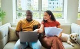 Two people sitting on a couch, one using a laptop and the other holding papers, with a small dog resting beside them.