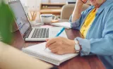 A person writing in a notebook while talking on the phone, with a laptop open on the desk and a coffee mug nearby.