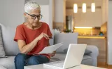 Older woman holding papers and working on laptop