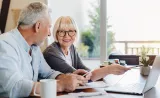 A senior couple reviews tax documents and reads from a laptop in their home office.