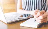 Woman writing notes looking at laptop and smartphone.